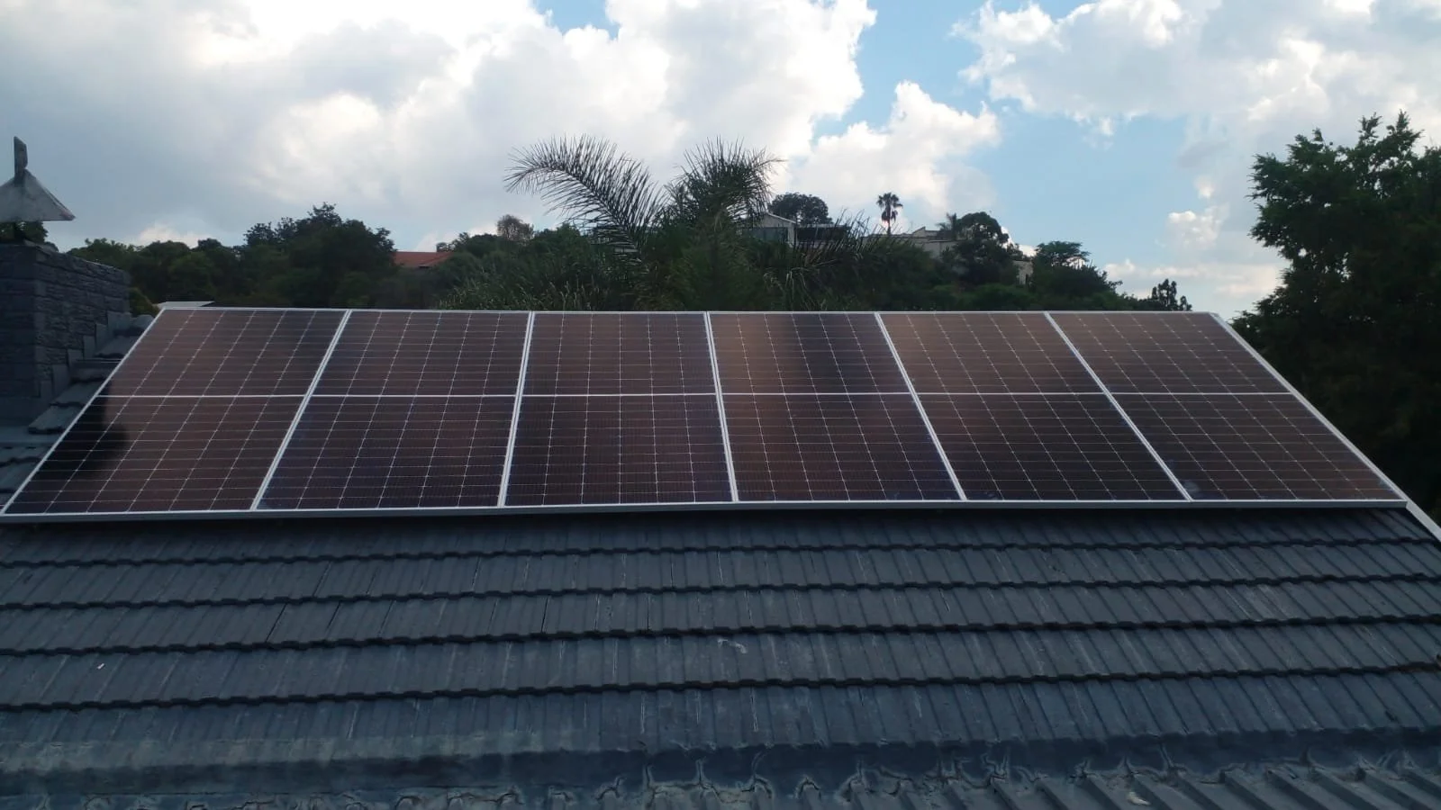 Photograph of solar panels installed on a roof, with trees and a partly cloudy sky in the background.