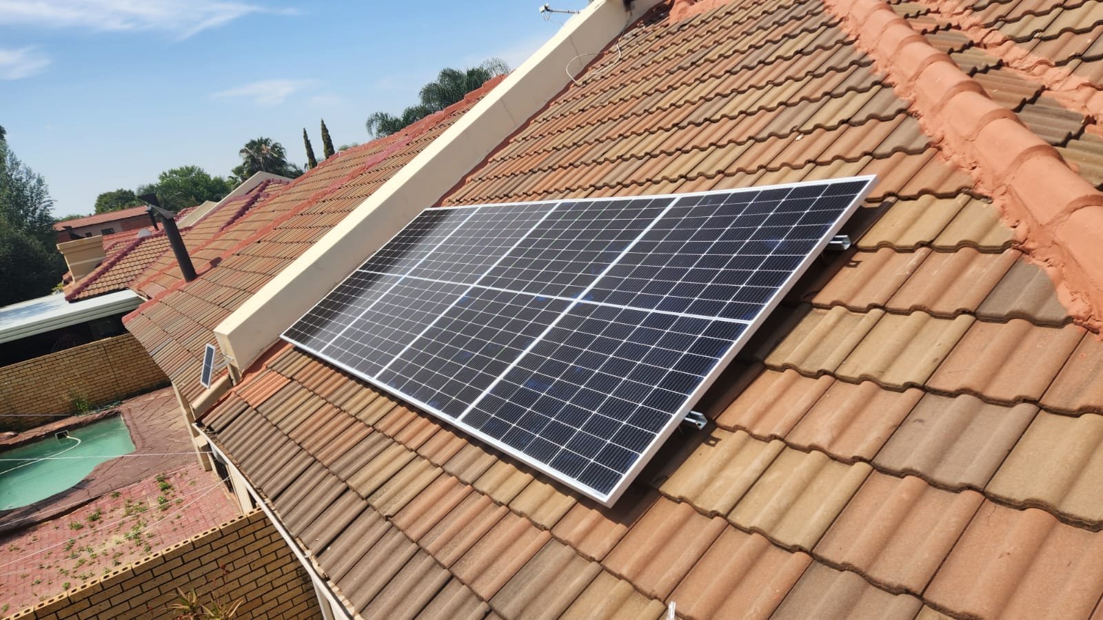 Photograph of solar panels installed on a sloped tiled roof of a house, with neighboring houses and trees visible in the background.
