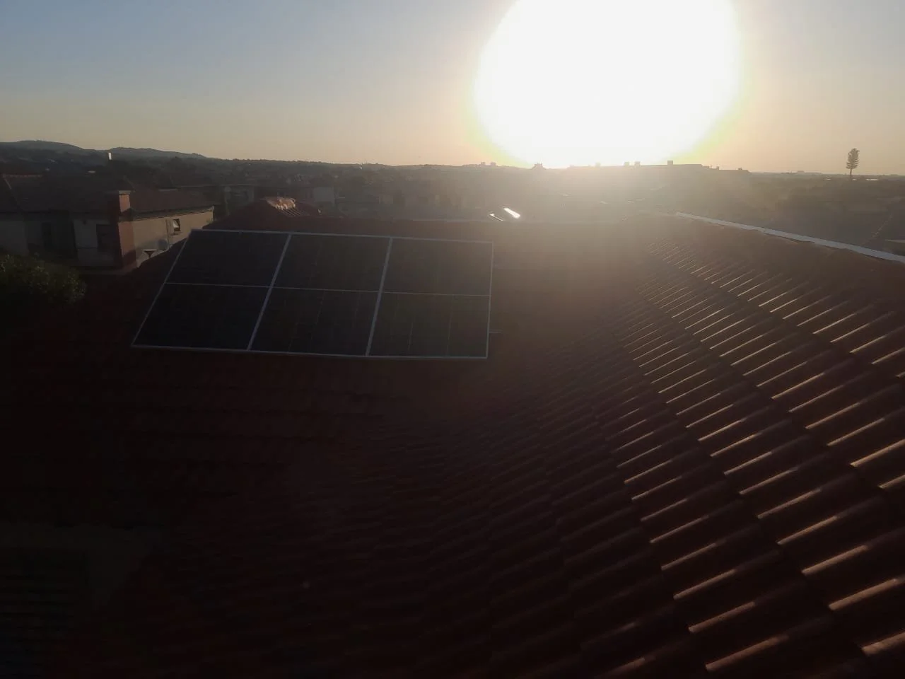 Sun setting on rooftops with solar panels on a tiled roof in a suburban area.