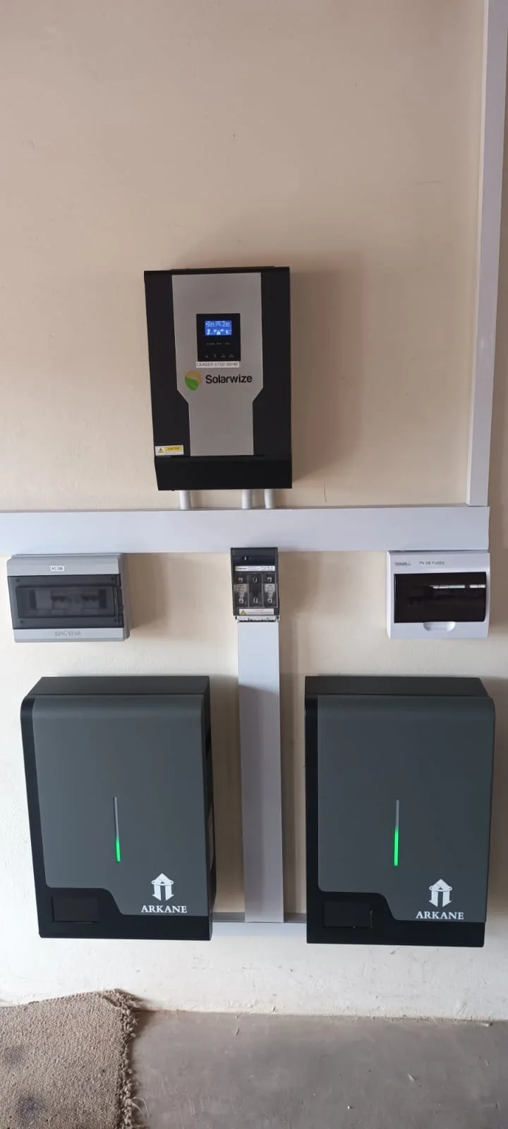 A solar power inverter system installed on a wall, with two gray batteries labeled 'ARKANE' below it, and a circuit breaker and a small electrical panel on either side.