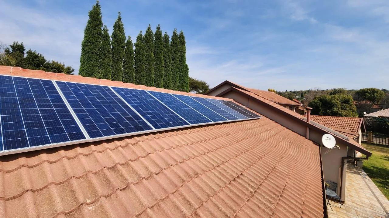 Solar panels installed on a house roof with red tiles, with several tall evergreen trees in the background and a partly cloudy blue sky.