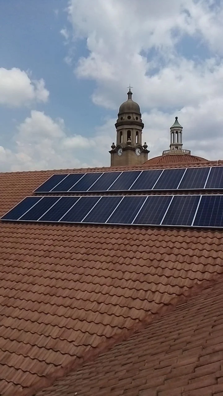Solar panels installed on a red-tiled roof with two church towers in the background under a partly cloudy sky.