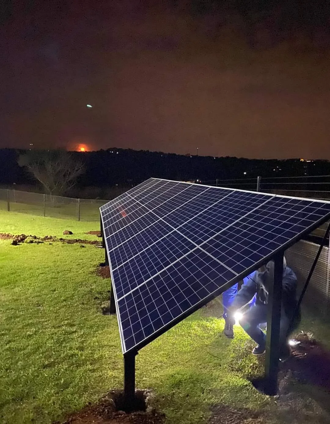 Nighttime view of a solar panel installation on a grassy area, with two workers crouching underneath it, illuminated by a flashlight. In the background, there is a dark landscape with the glow of a distant fire or explosion on the horizon and a faint