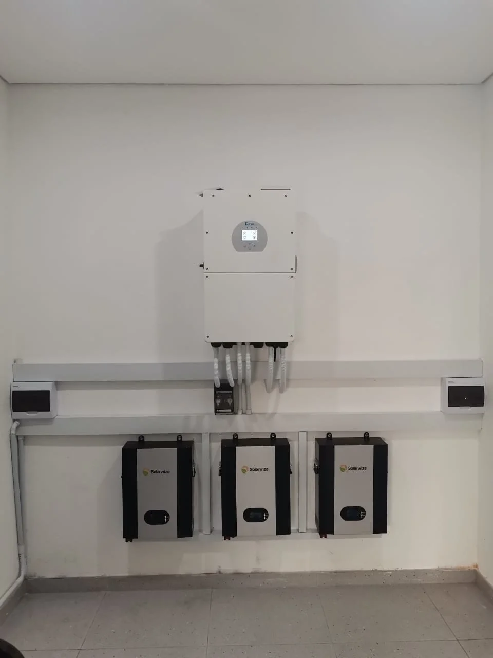 Wall-mounted solar inverter system with three inverters labeled 'Solarwize', a larger central inverter with digital display, and multiple cables connected downward, set against a plain white wall in an indoor setting.