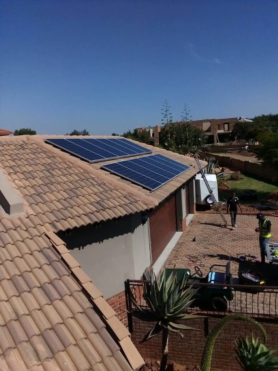 Residential house with solar panels installed on the tiled roof, two workers near equipment in the driveway, and desert plants in the yard under a clear blue sky.