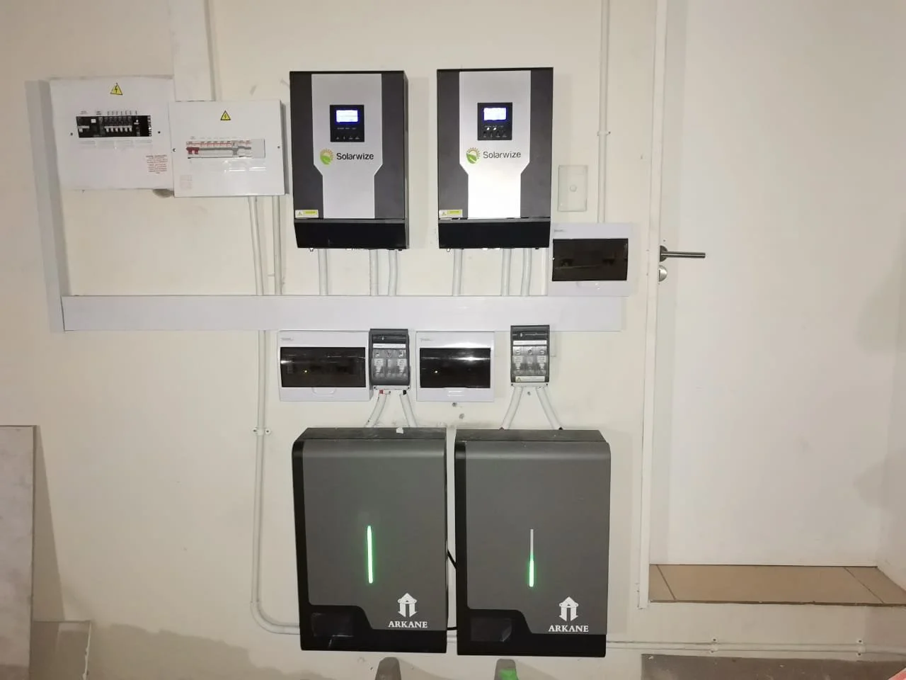 Electrical control panel and inverters installed on a wall, with wires connected, including two Solarwize inverters, two smaller controllers, and large gray battery units labeled 'ARKANE' at the bottom.