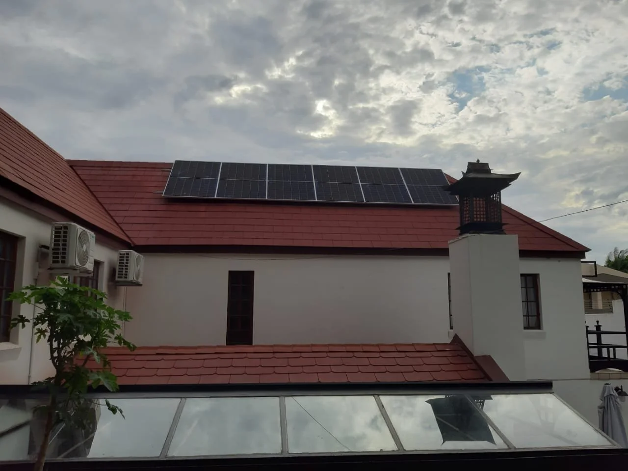 House with a red-tiled roof, solar panels on top, and chimneys, under a cloudy sky.