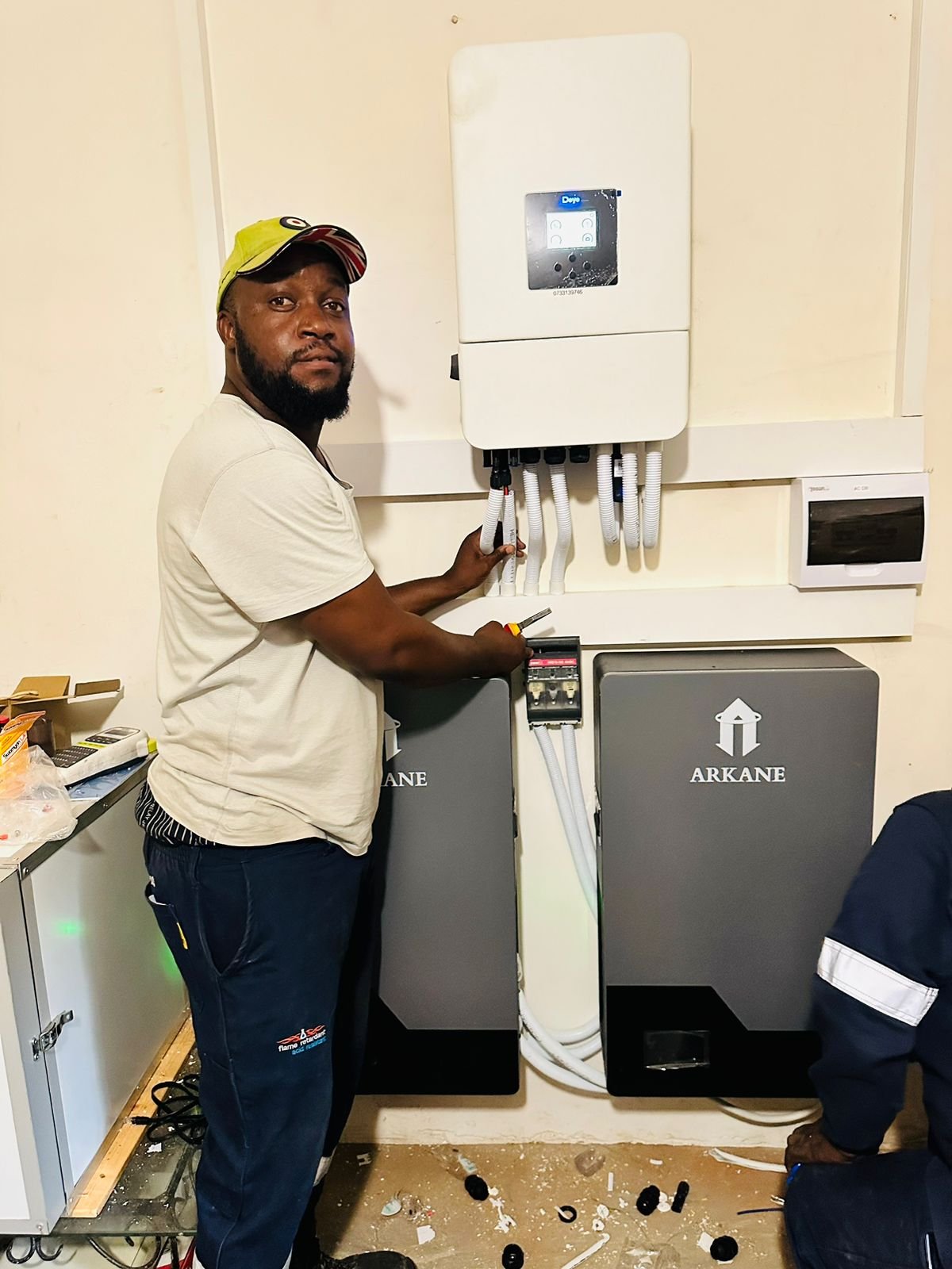 A man working on electrical or solar equipment, standing next to a control panel and two Arkane units, with tools in hand, in a room with a cluttered floor.