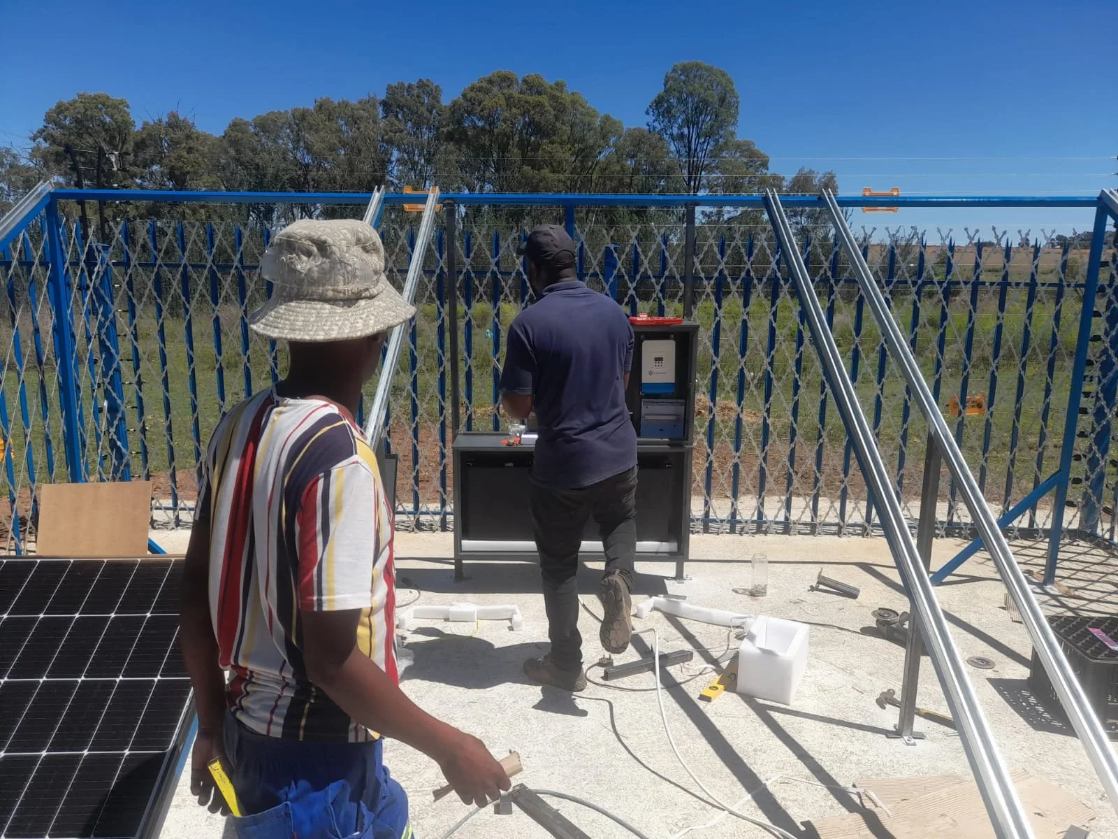 Two men working on solar panel installation outdoors on a sunny day, with a metal fence and trees in the background.