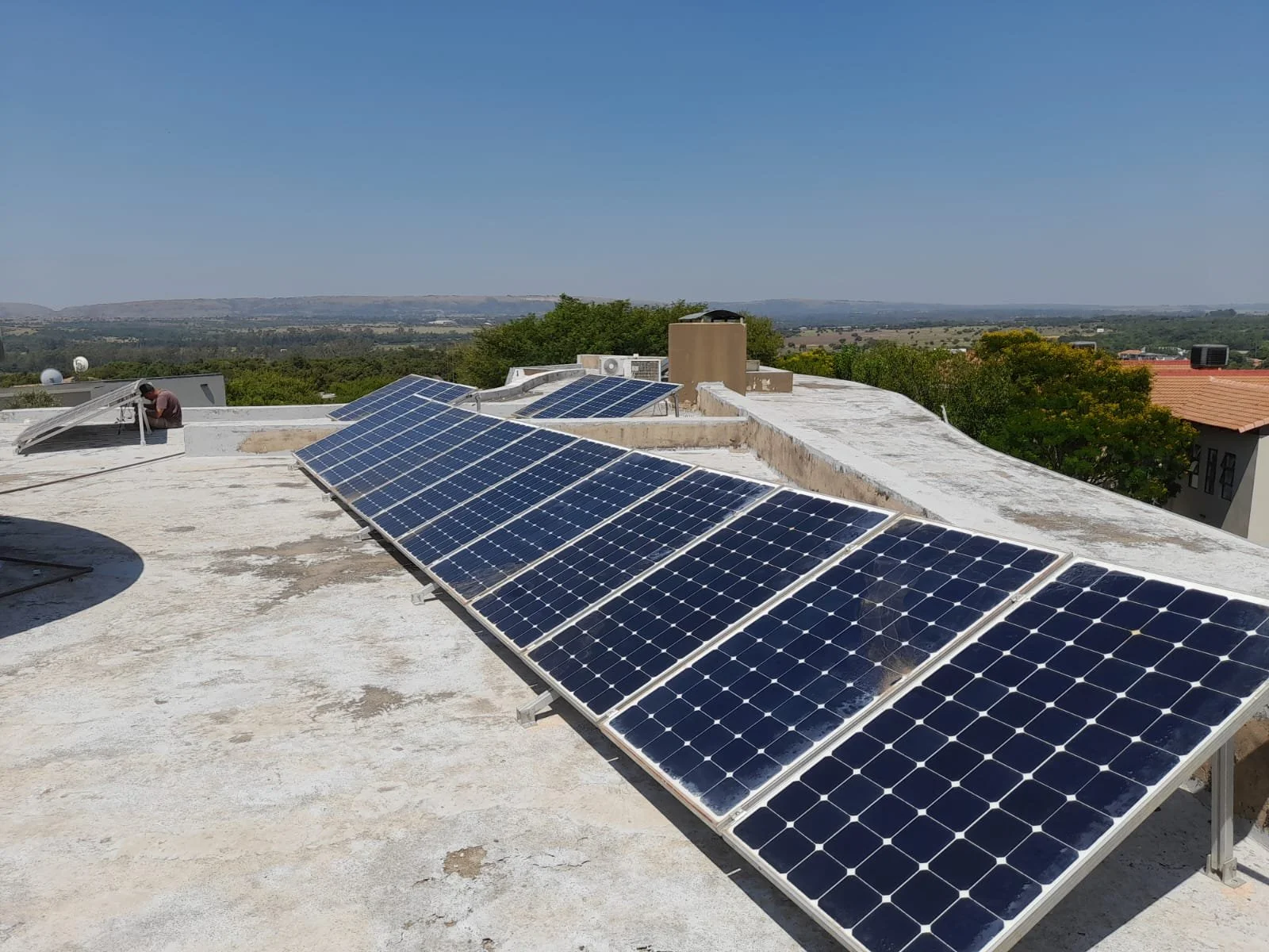 On a flat rooftop, multiple blue solar panels are installed, with one person working near another panel. The background shows trees and a distant landscape under a clear blue sky.