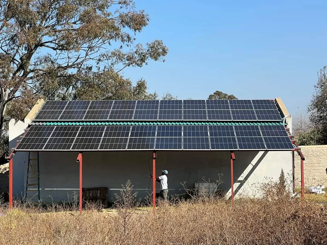 A building with solar panels installed on its roof, supported by red metal beams. A person wearing a white shirt and blue cap is working in front of the building. There are trees and dry grass in the background.
