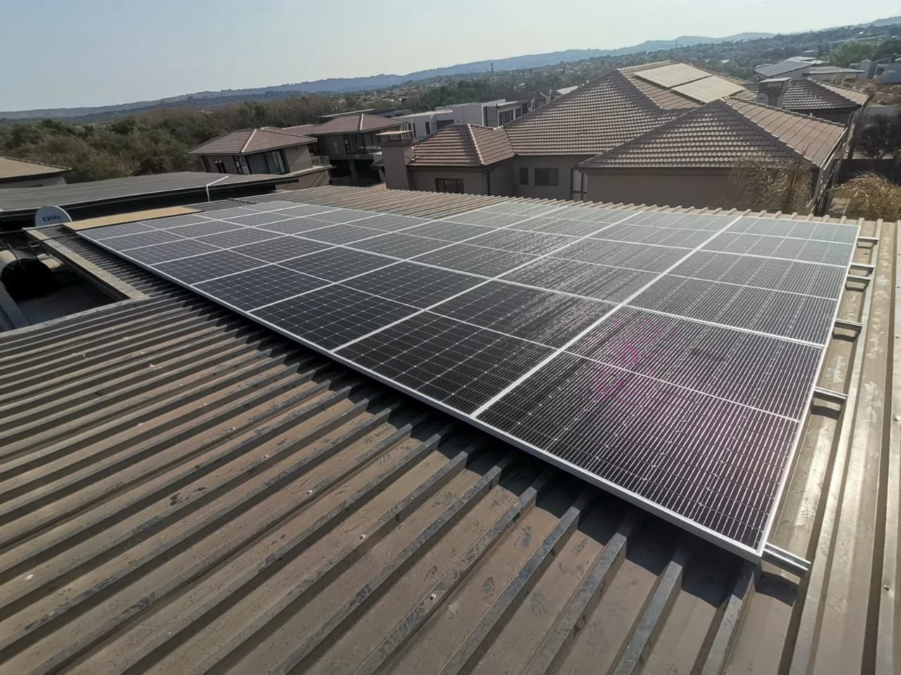 Solar panels installed on a rooftop with neighboring houses and hilly landscape in the background.