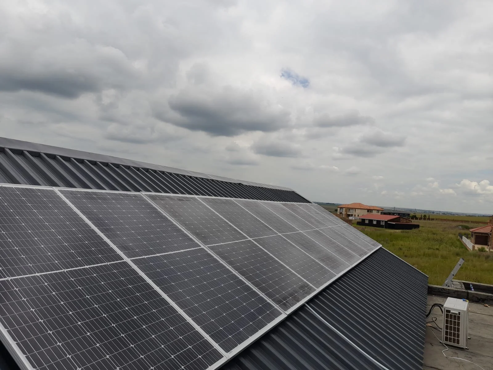 Solar panels installed on the roof of a building, with a cloudy sky and distant houses in the background.