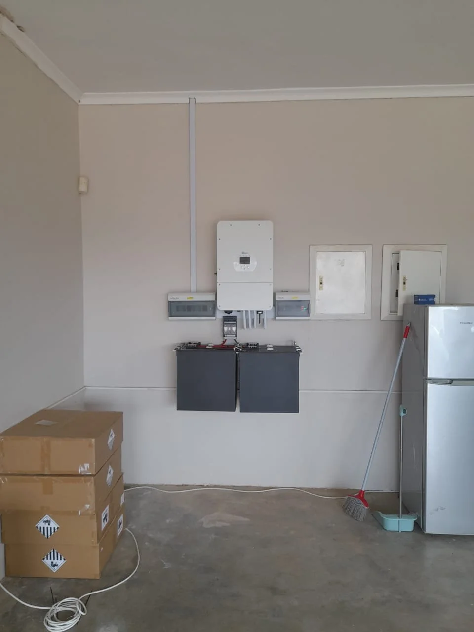 Empty garage with three cardboard boxes, a broom and dustpan, a refrigerator, and electrical panels on the wall.