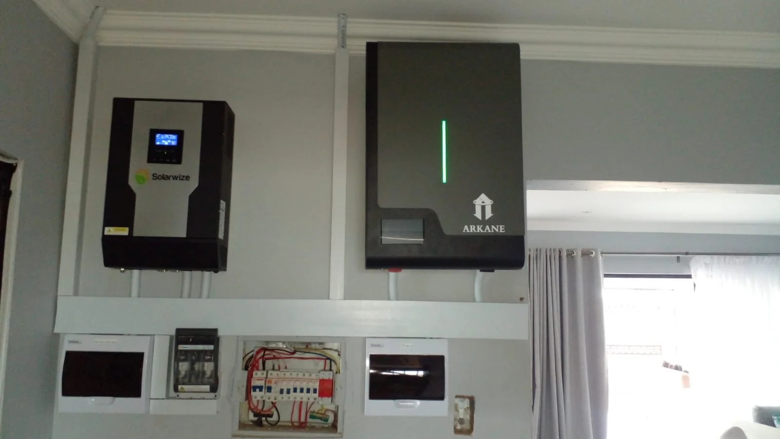 Electrical utility equipment on a wall, including a solar inverter labeled 'Solarwize' on the left, a large gray battery with a green LED light and the logo 'ARKANE' on the right, and an electrical panel with circuit breakers and wiring below.