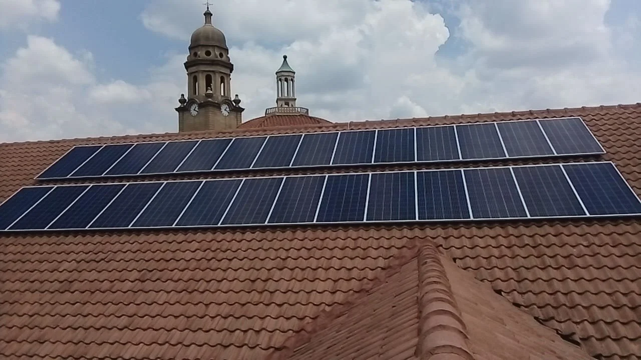 Solar panels installed on a red tiled roof with a church steeple and clouds in the background.