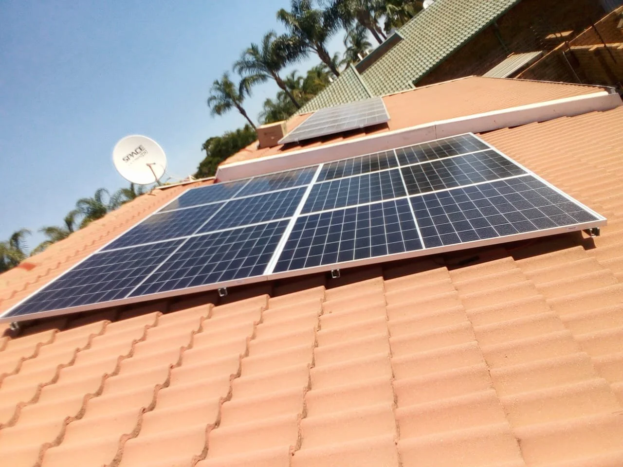 Solar panels installed on a terracotta tile roof with a satellite dish and palm trees in the background under a clear blue sky.