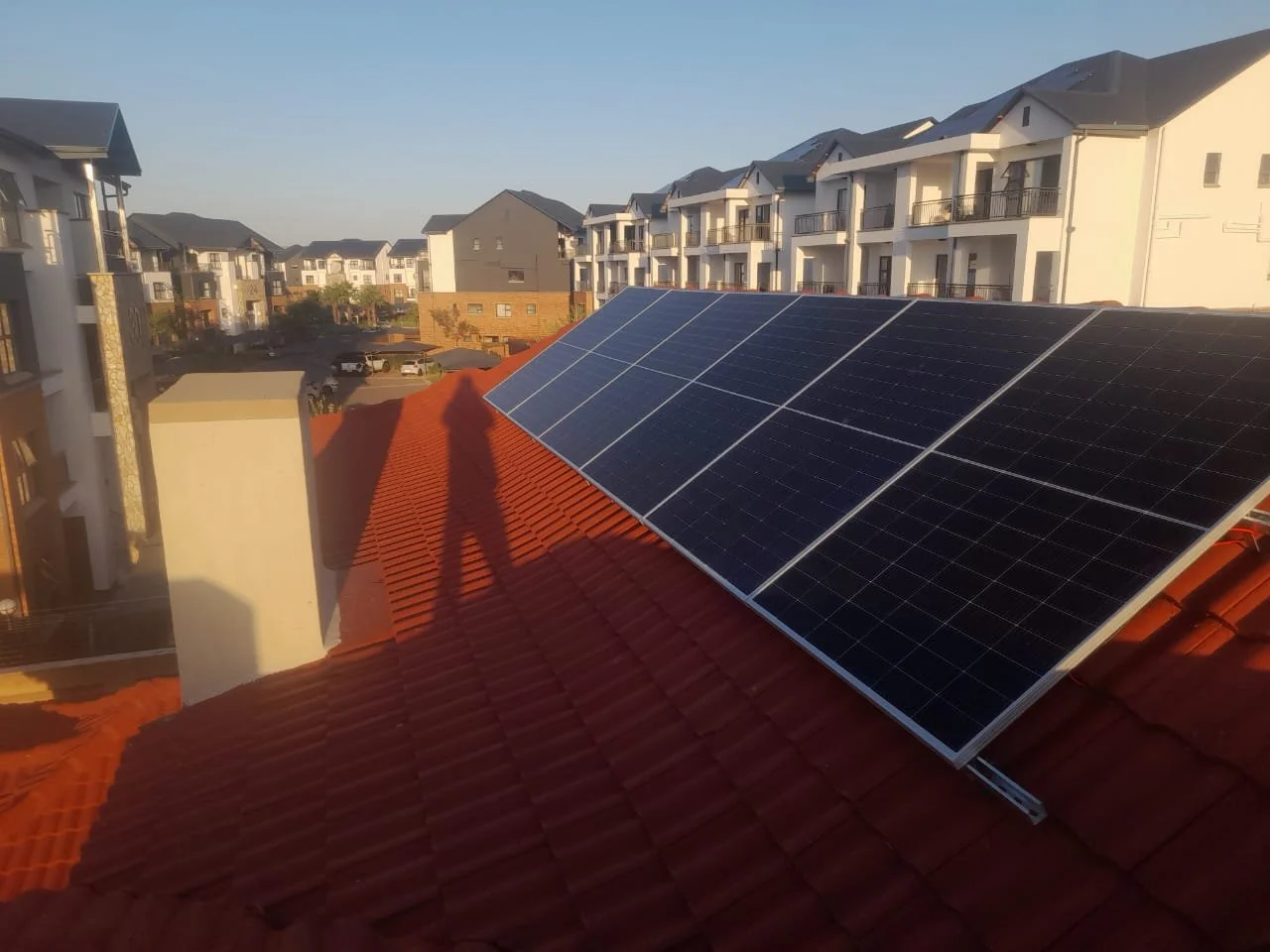 Solar panels installed on a red-tiled roof of a residential building, with a view of nearby white and gray apartment buildings and a parking lot in the background.