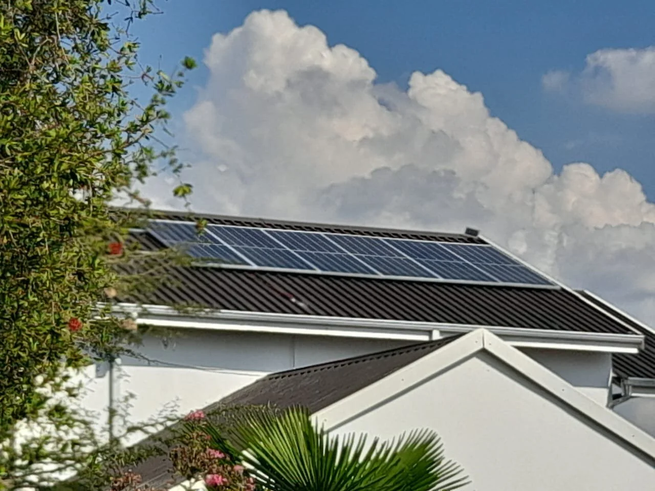A house with solar panels on the roof, surrounded by trees and bushes, under a partly cloudy sky.
