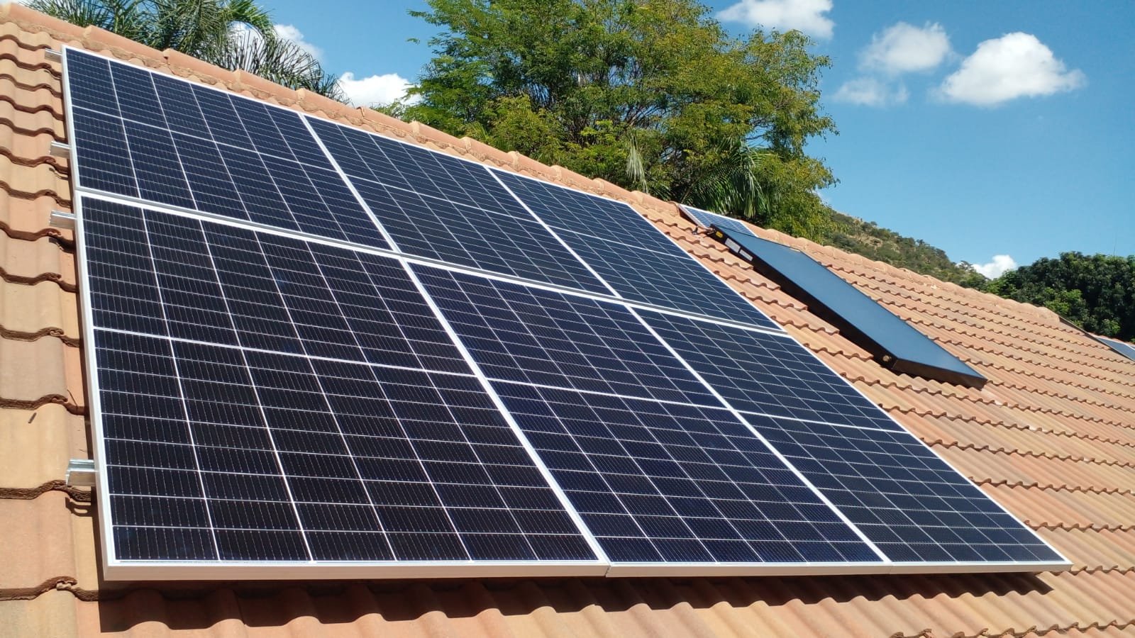 Solar panels installed on a terracotta tiled roof with trees and a blue sky in the background.