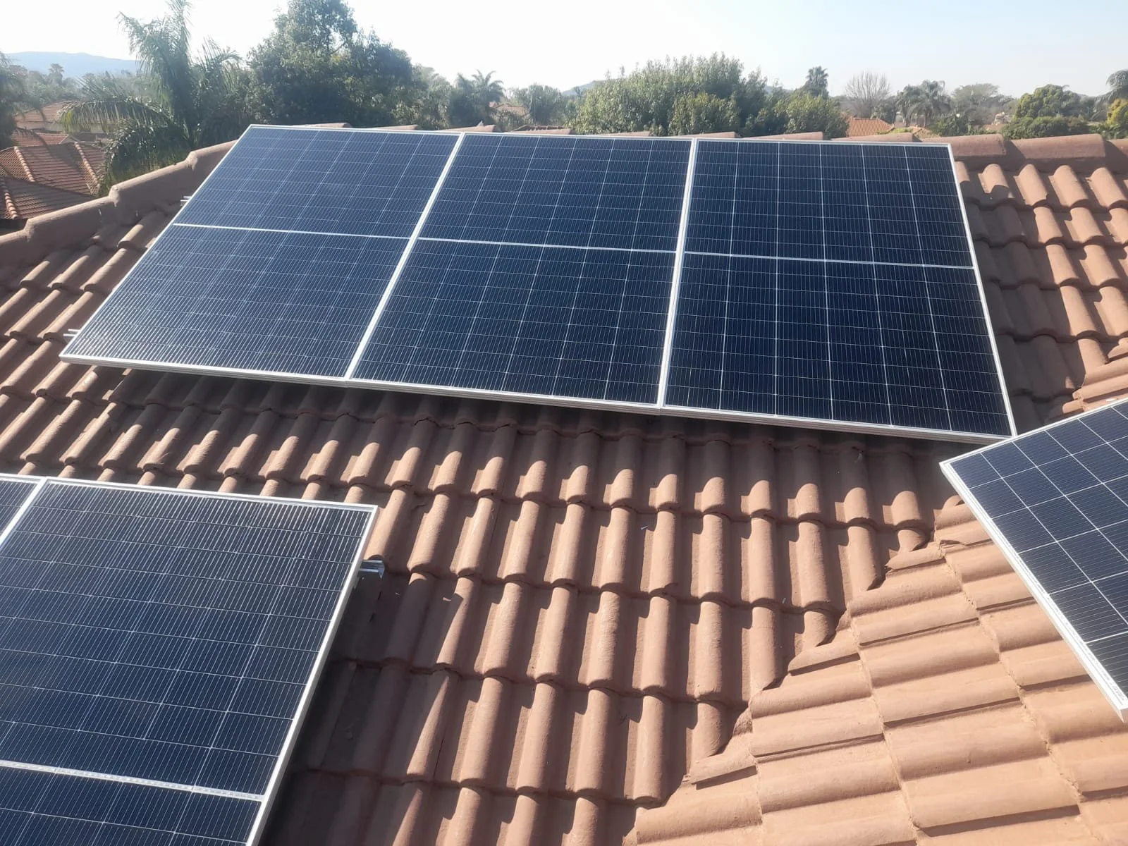 Multiple blue solar panels installed on a brown tiled rooftop with trees and houses in the background.