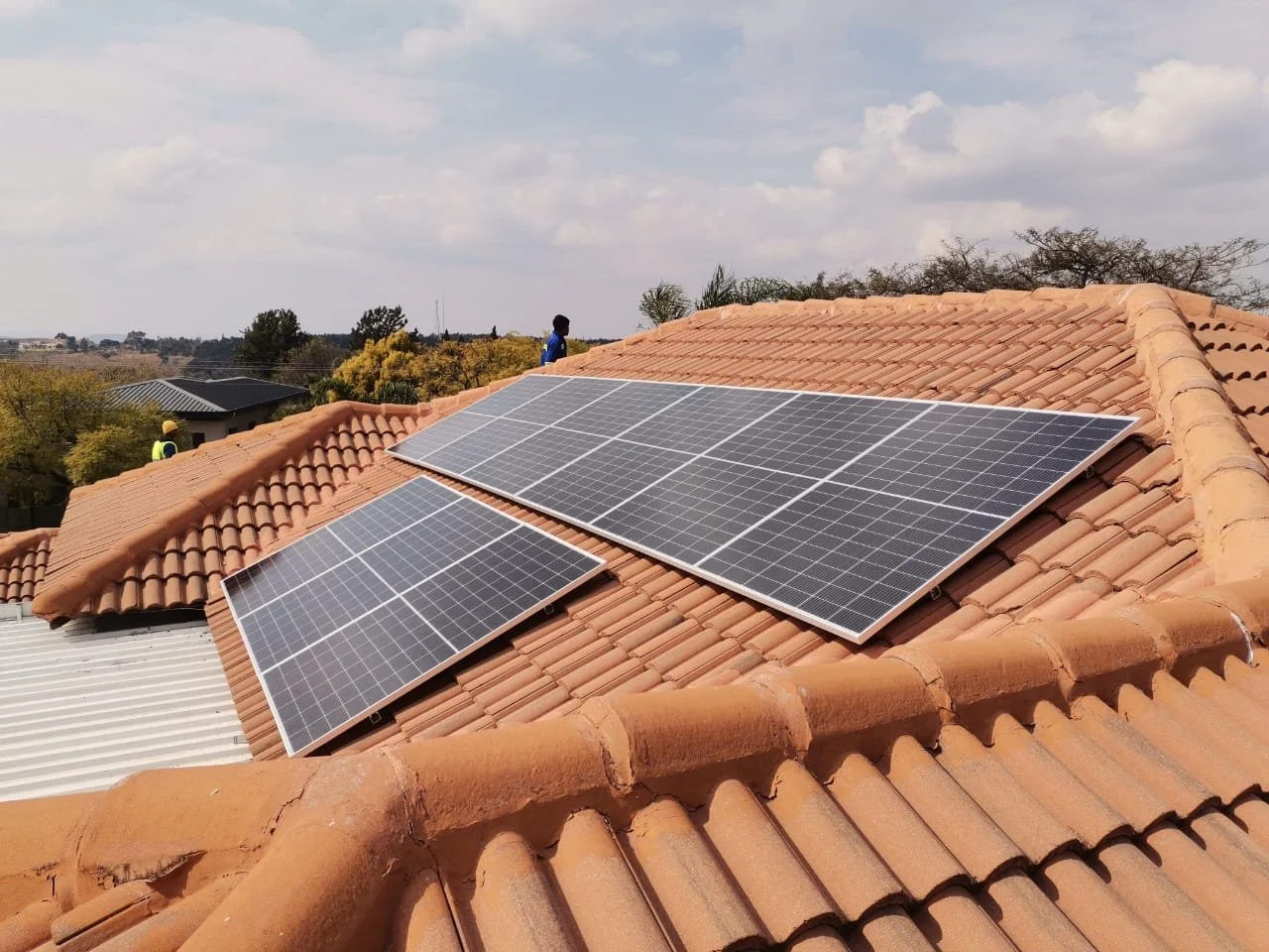 Solar panels installed on a terracotta tile roof with a cloudy sky and trees in the background.
