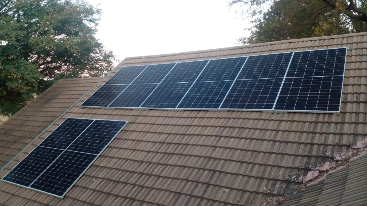 Multiple solar panels installed on a sloped roof with brown tiles, with trees in the background.