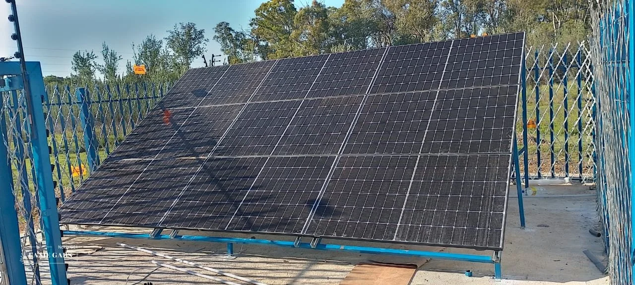 Photograph of a solar panel installed at a rooftop, enclosed by blue fencing, with trees and blue sky in the background.