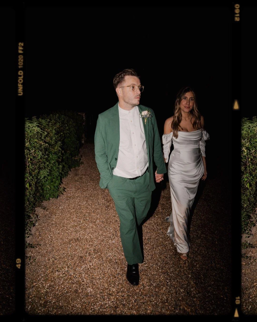Wedding ceremony photo of a man and woman walking hand in hand outdoors at night, the groom in a green suit with a white shirt and a boutonniere, the bride in a satin off-shoulder gown.