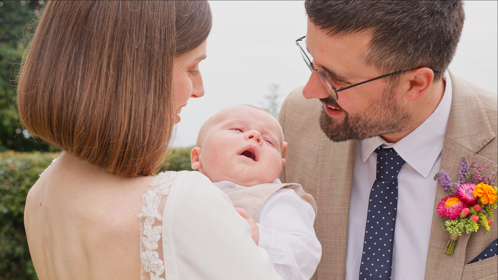 At a wedding a bride with brown hair holding a baby, with a groom in glasses and a beige suit with a boutonniere, smiling at the baby outdoors after the ceremony.