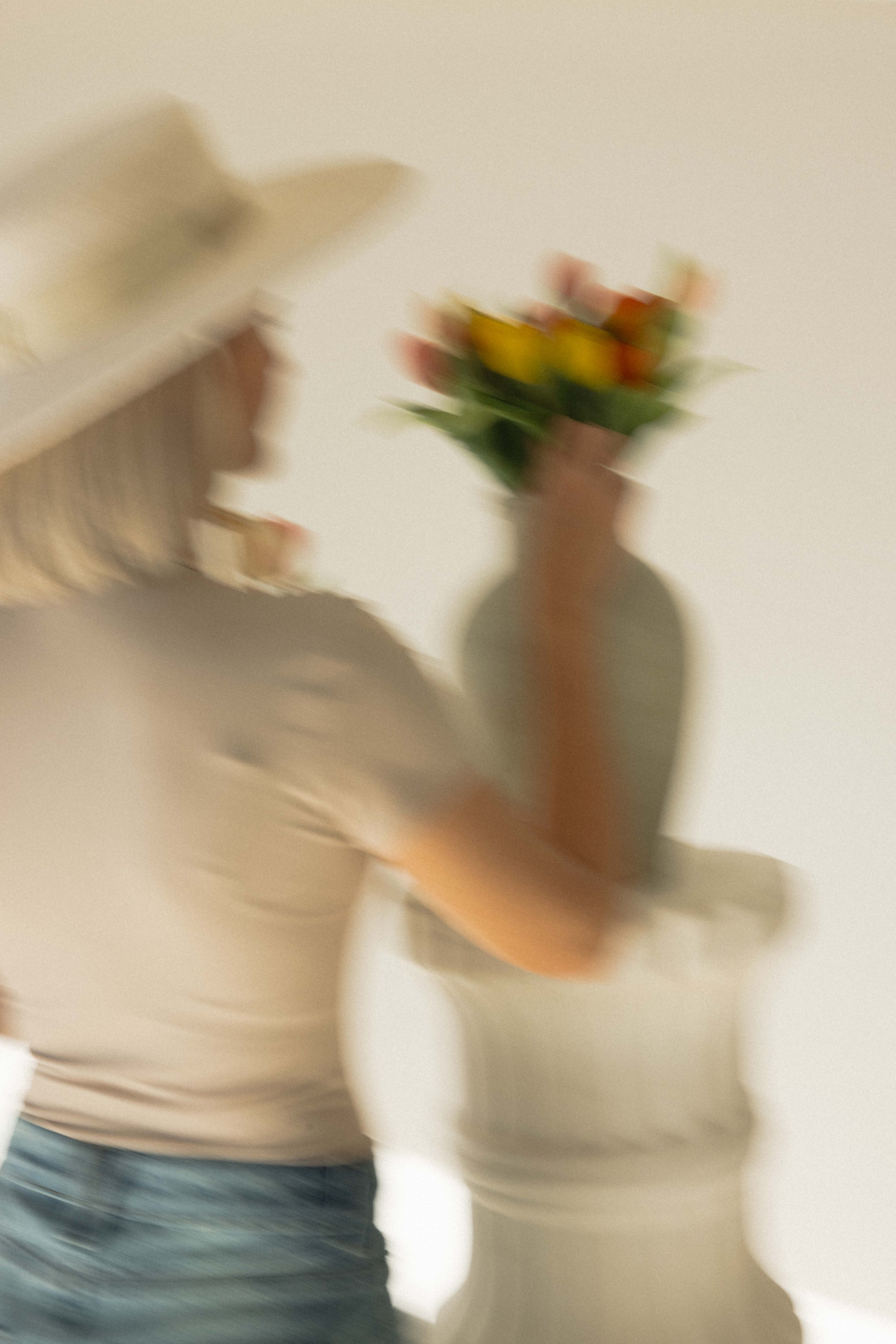A woman arranging a bouquet of colourful faux silk flowers, wearing a beige long-sleeve shirt and jeans, with a white plinth in the background on which the flowers are displayed in a flower pot in Standerson, South Africa.