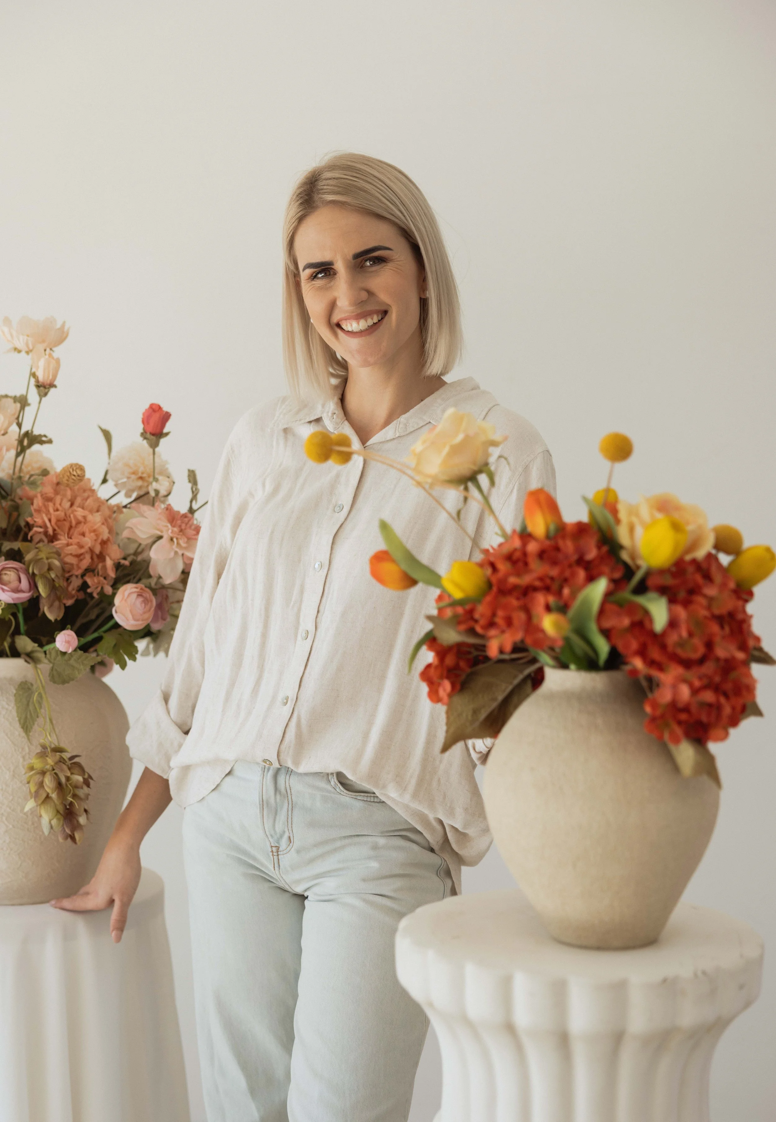 A woman with blonde hair smiling, standing between two large vases filled with colourful silk flowers, wearing a light-colored shirt and pants, against a plain white background.