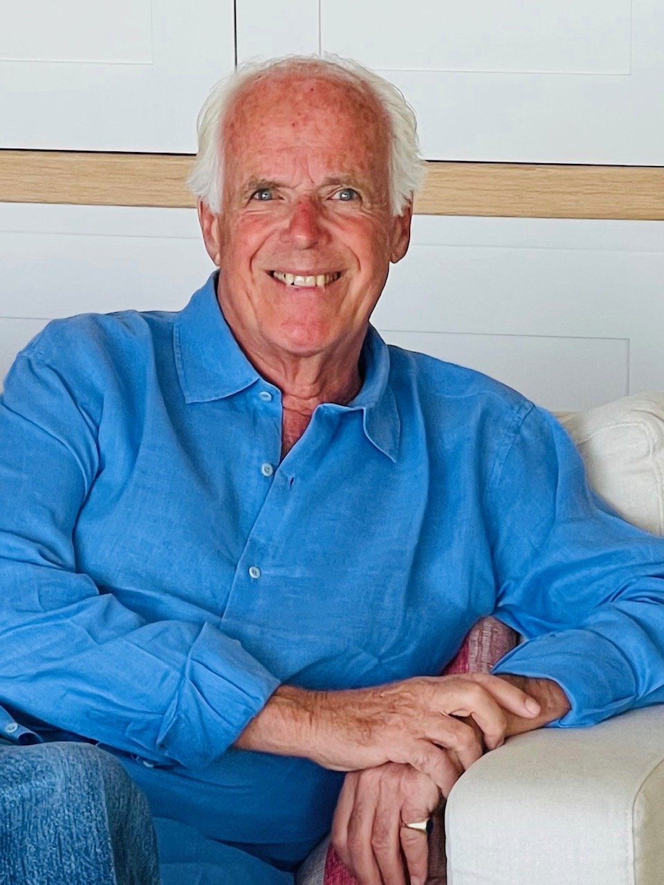 Smiling elderly man with white hair and a blue shirt sitting on a beige couch indoors.