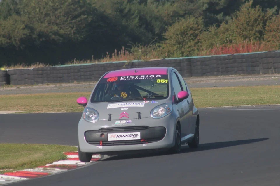 A small racing car on a track, gray with pink side mirrors, nose, and windshield banner, navigating a curve while leaning into the turn.