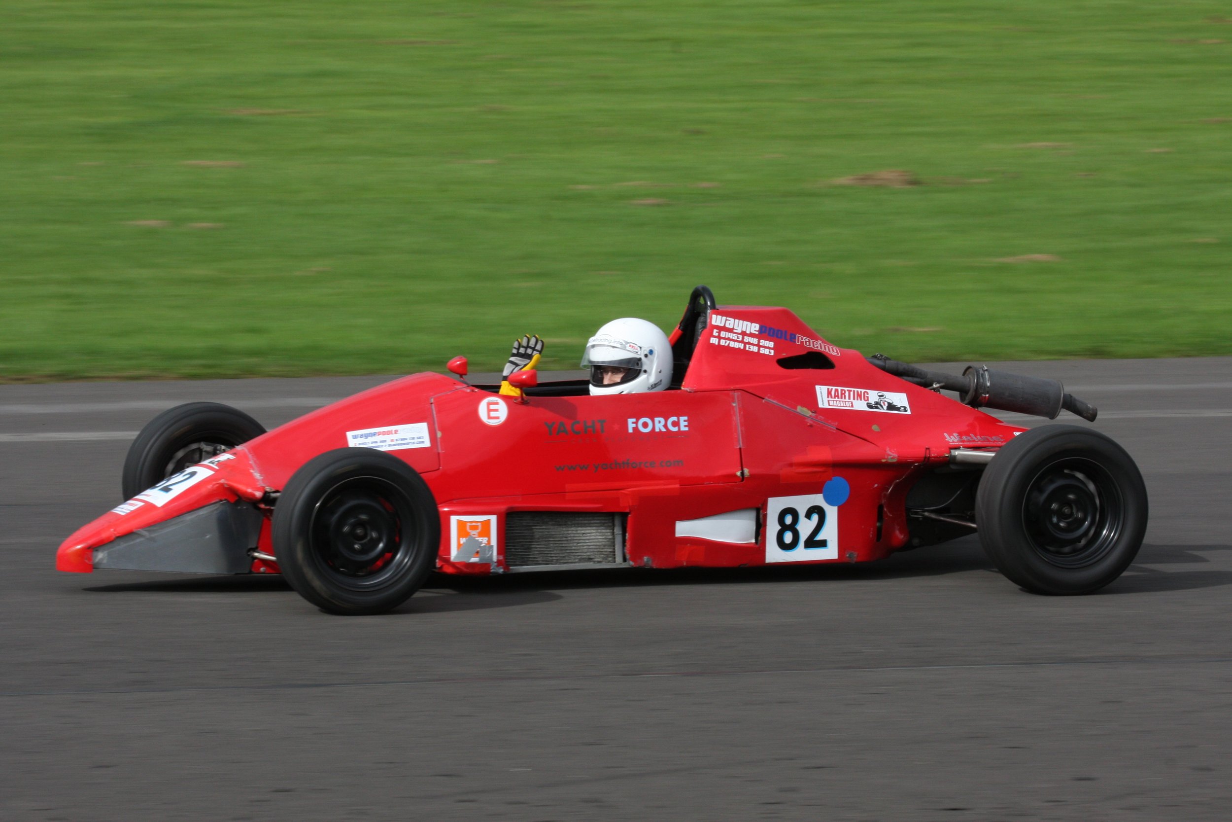 Red race car with the driver in a white helmet and yellow gloves, driving on a racetrack with green grass background.