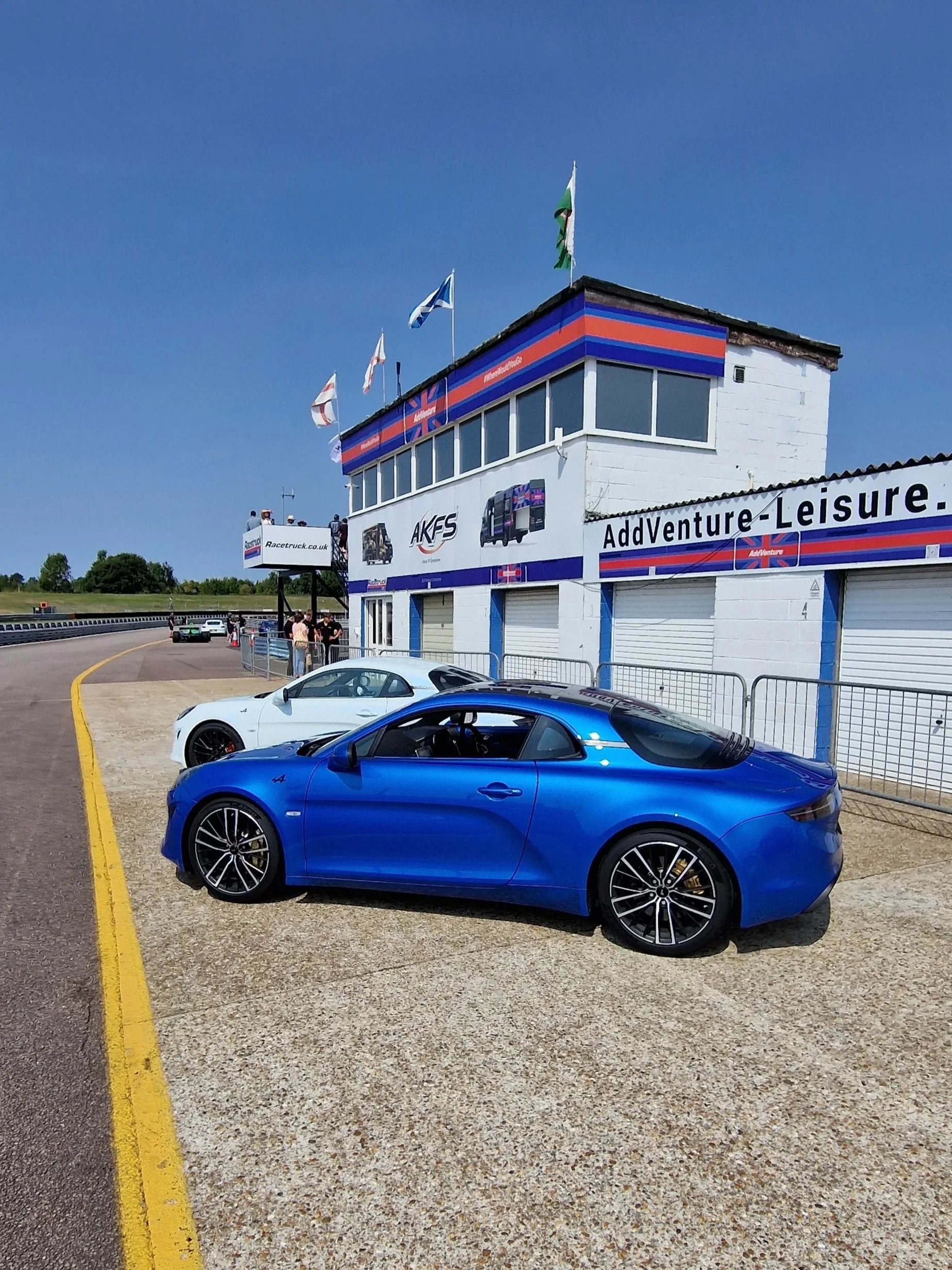 A blue sports car and a white sports car parked at a race track paddock area with a building in the background displaying flags, advertisements