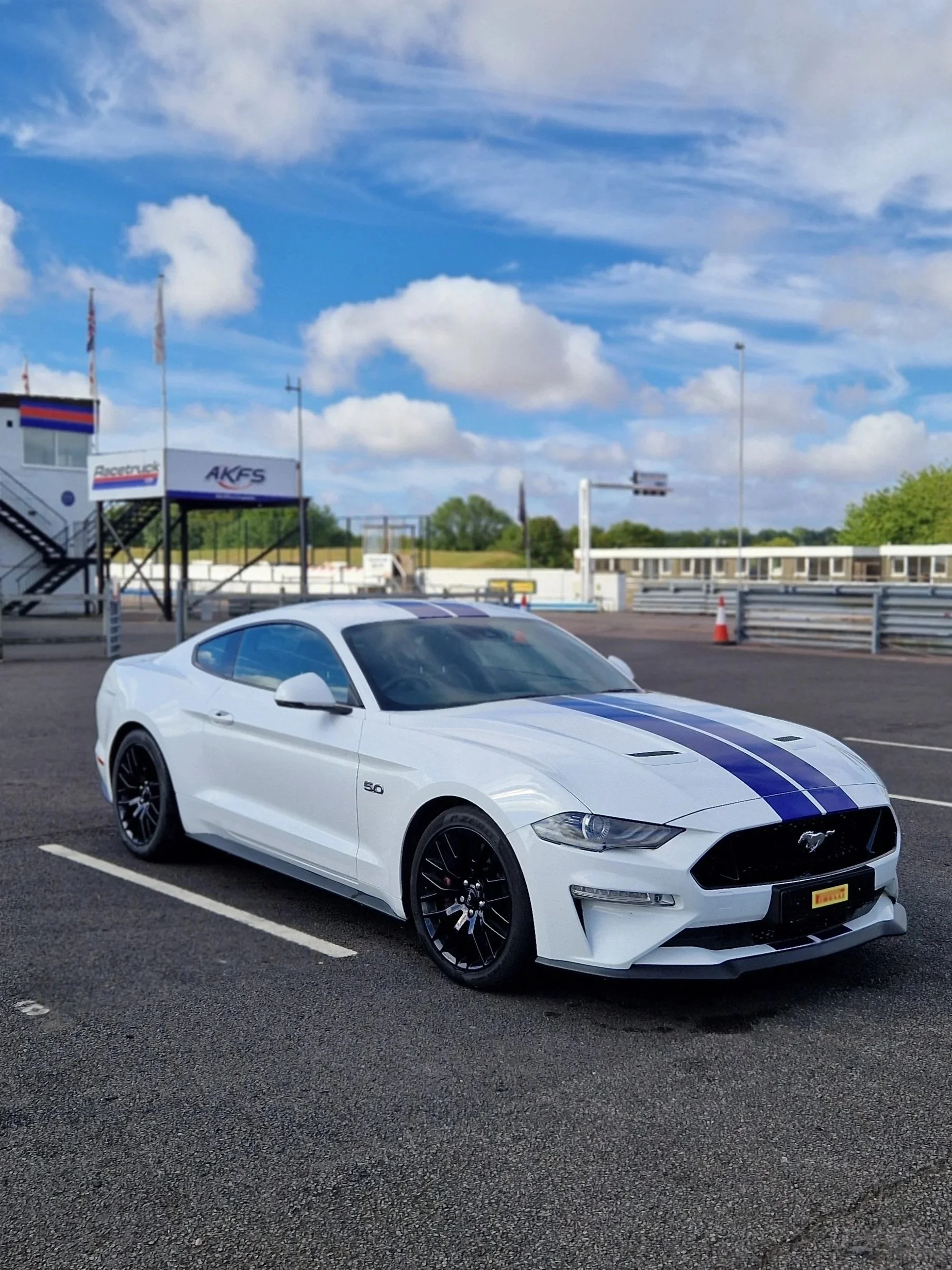 A white Ford Mustang sports car with blue racing stripes parked on asphalt at a race track. The sky is partly cloudy with patches of blue, and there are some structures and orange traffic cones in the background.
