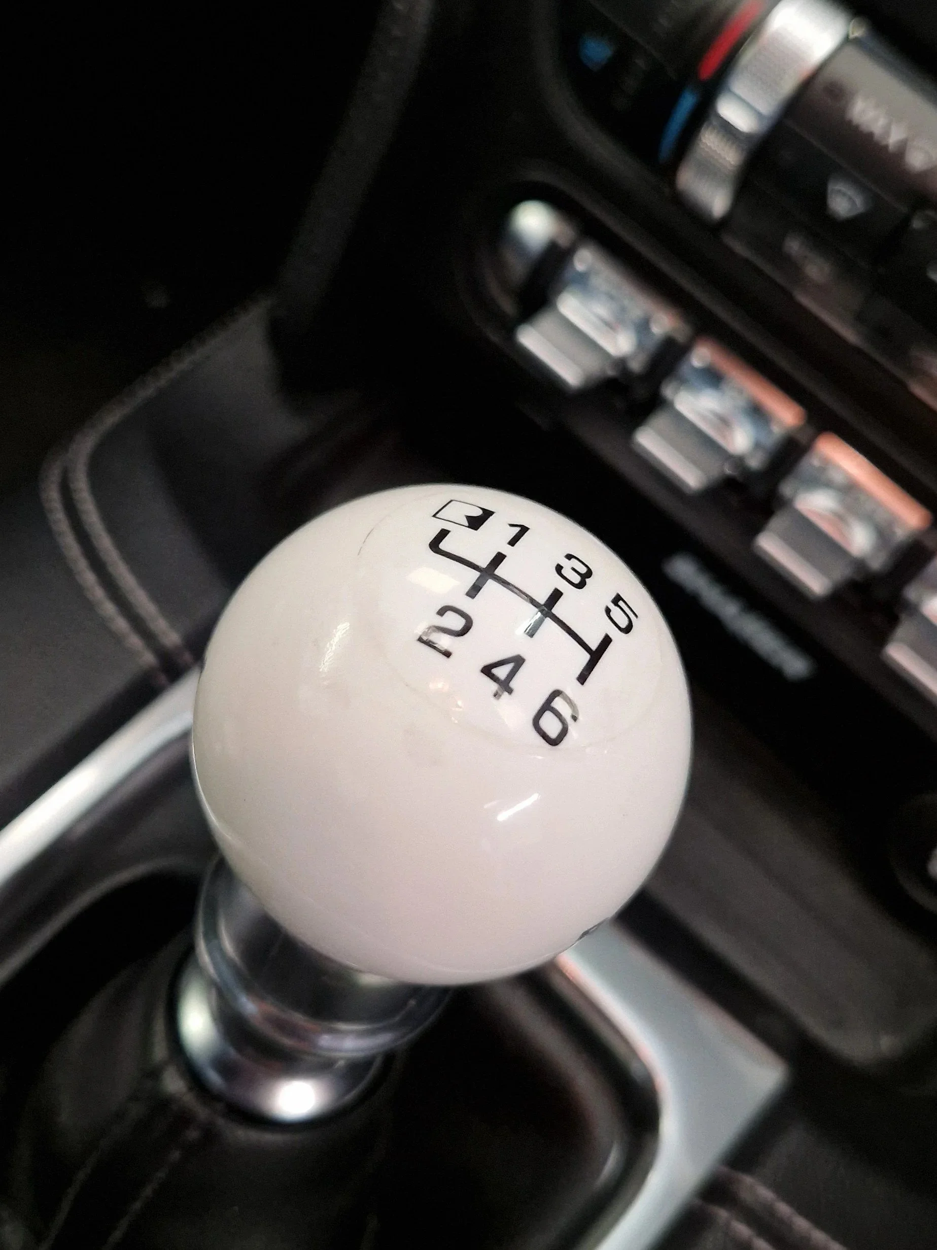 A close-up of a car's manual gear shift knob with a white top showing a 6-speed pattern and reverse, situated in the center console of a car interior with black leather and metallic accents.