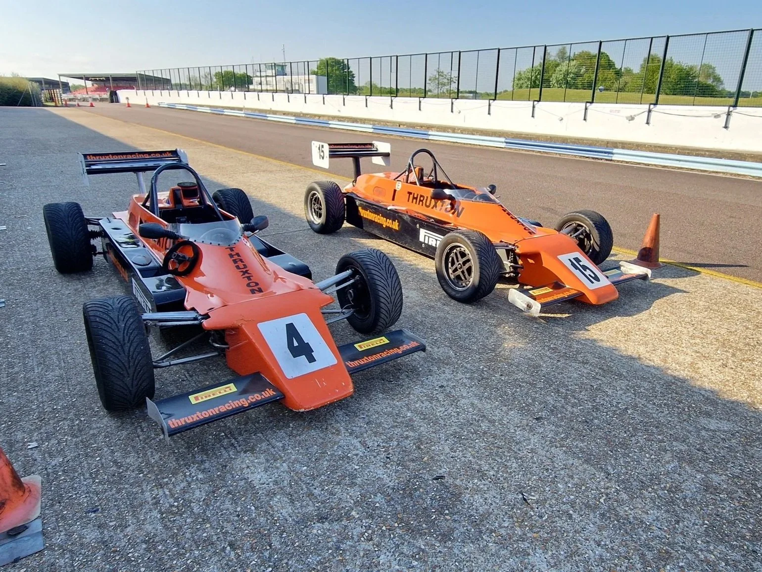 Two orange race cars parked on a race track's pit area, with traffic cones and metal barriers on the side. The cars have the number 4 and 15.