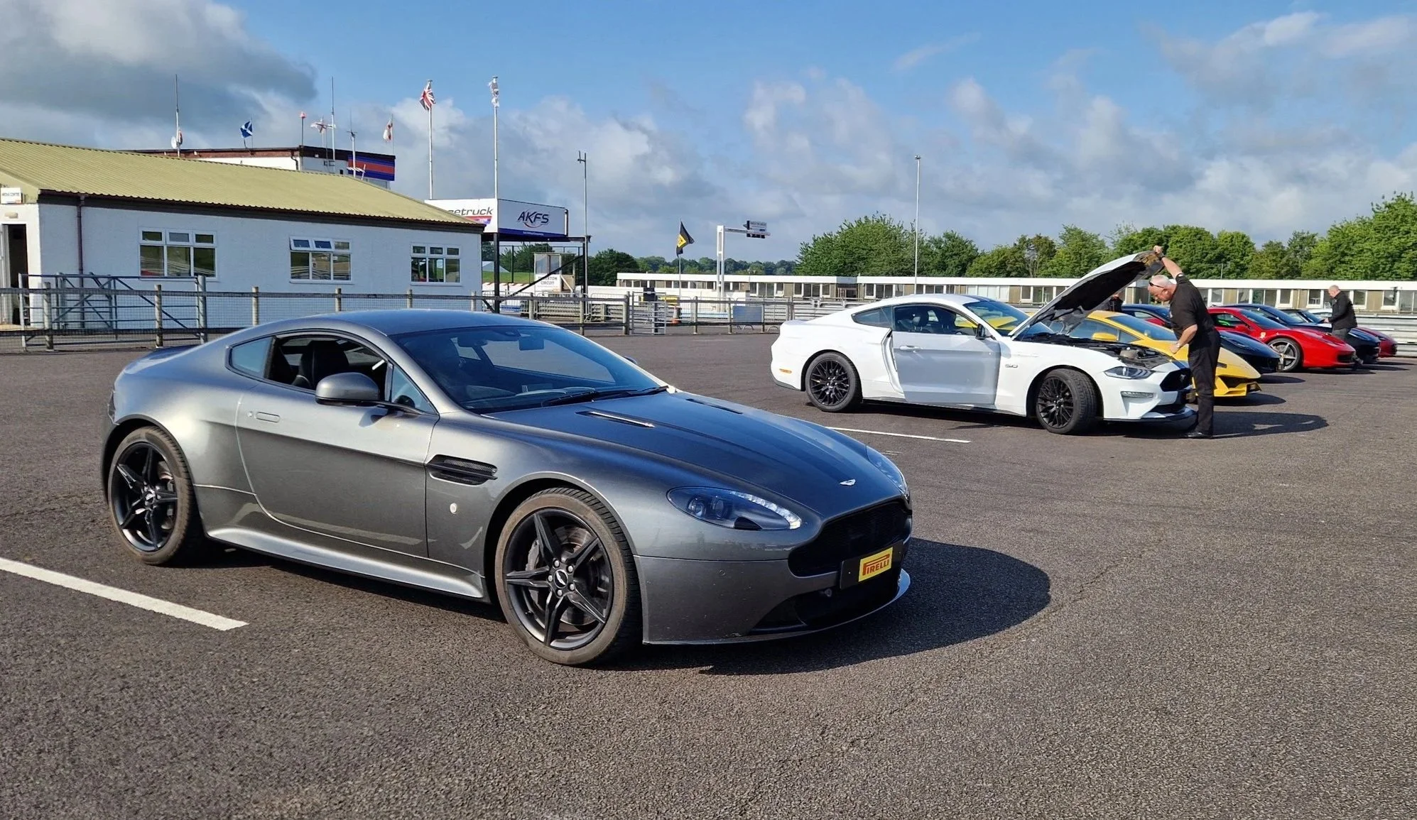 A row of sports cars parked in a lot with two men inspecting one of the cars, which has its hood open. The cars include a gray Aston Martin and a white modern Mustang. The lot has a building and flags in the background under a partly cloudy sky.