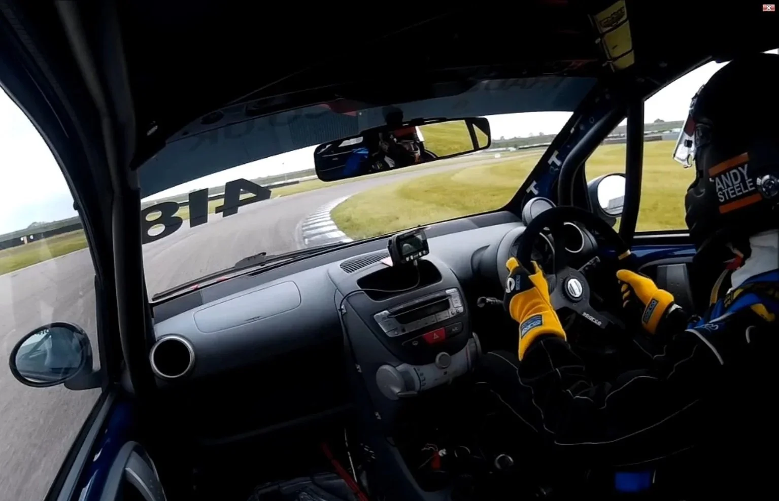 The interior view of a race car shows a driver wearing a helmet and racing suit, gripping the steering wheel, with a curving race track visible through the windshield.