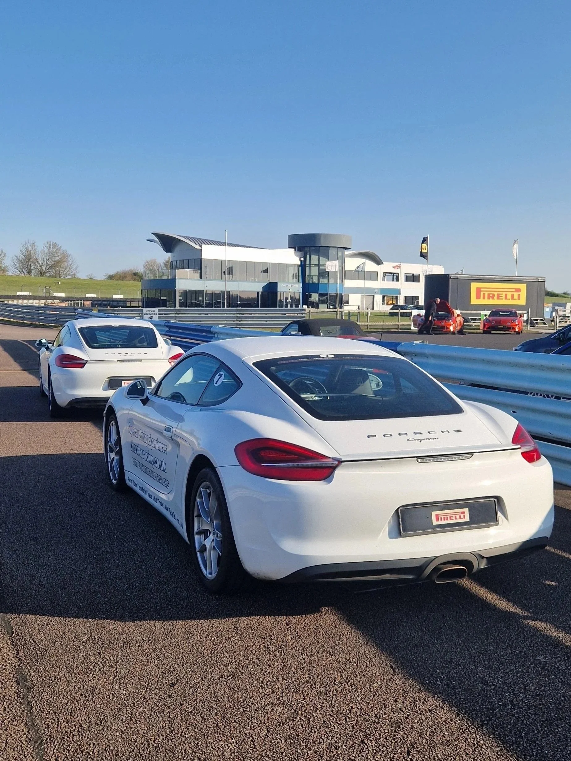 White Porsche sports car parked on a race track under a clear blue sky, with a modern building and other cars in the background.