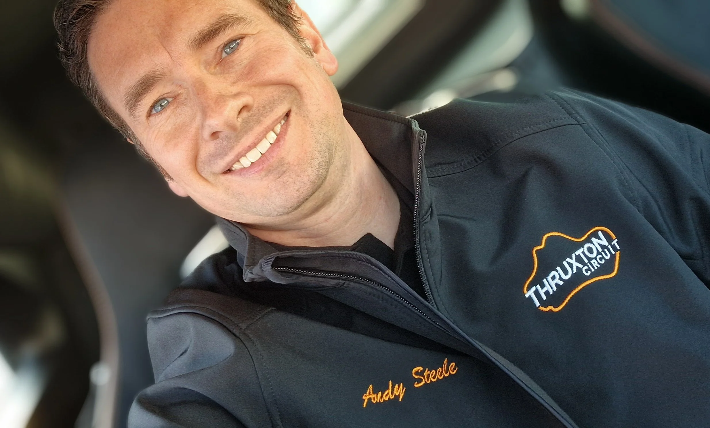 Andy Steele smiling and wearing a black jacket with the logos 'Thruxton Circuit' and ‘Andy Steale’ embroidered on it.
