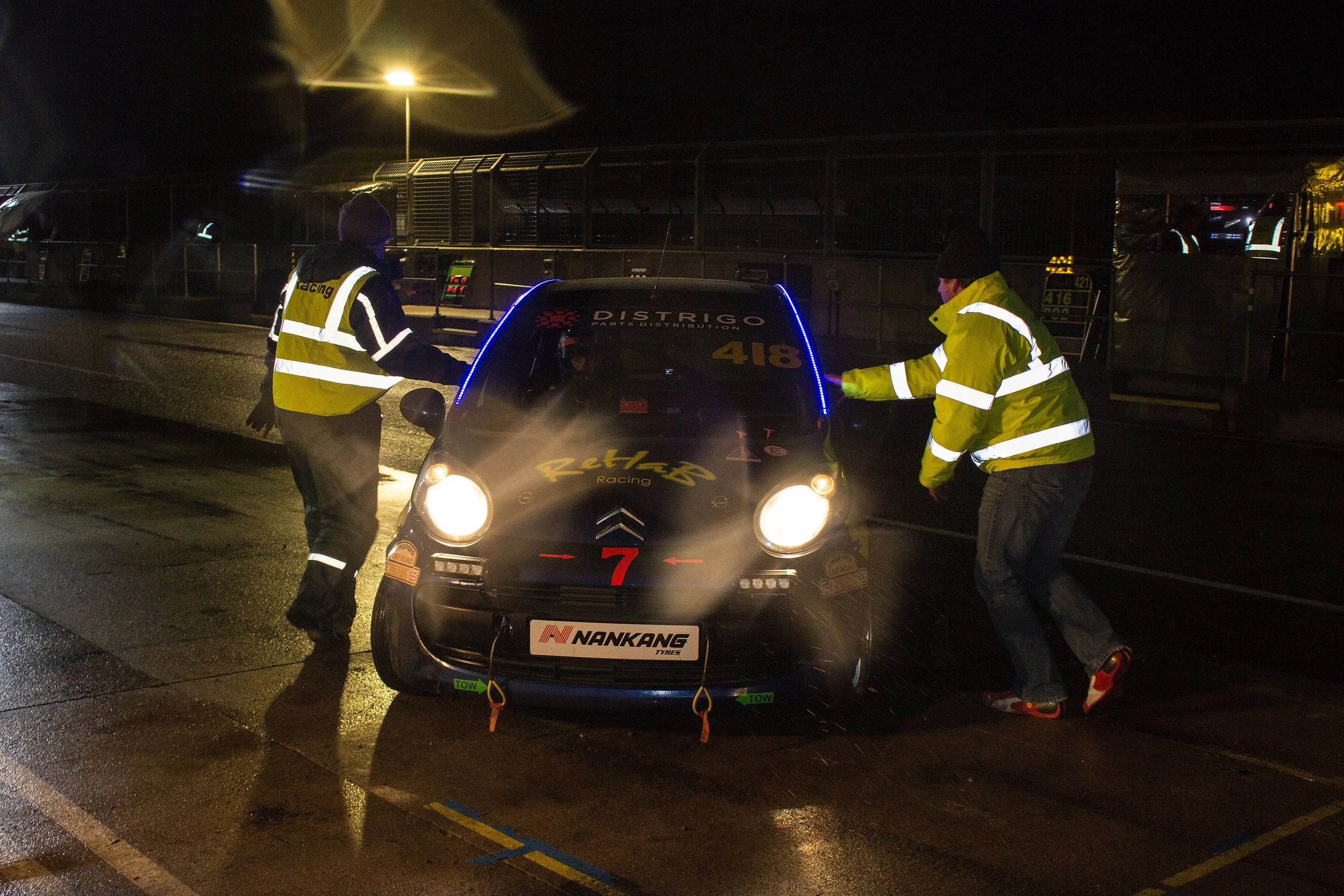 Two race car mechanics or officials working on a black Citroën C1 race car in the rain at night, wearing yellow and black reflective jackets, with bright headlights and rain droplets visible.