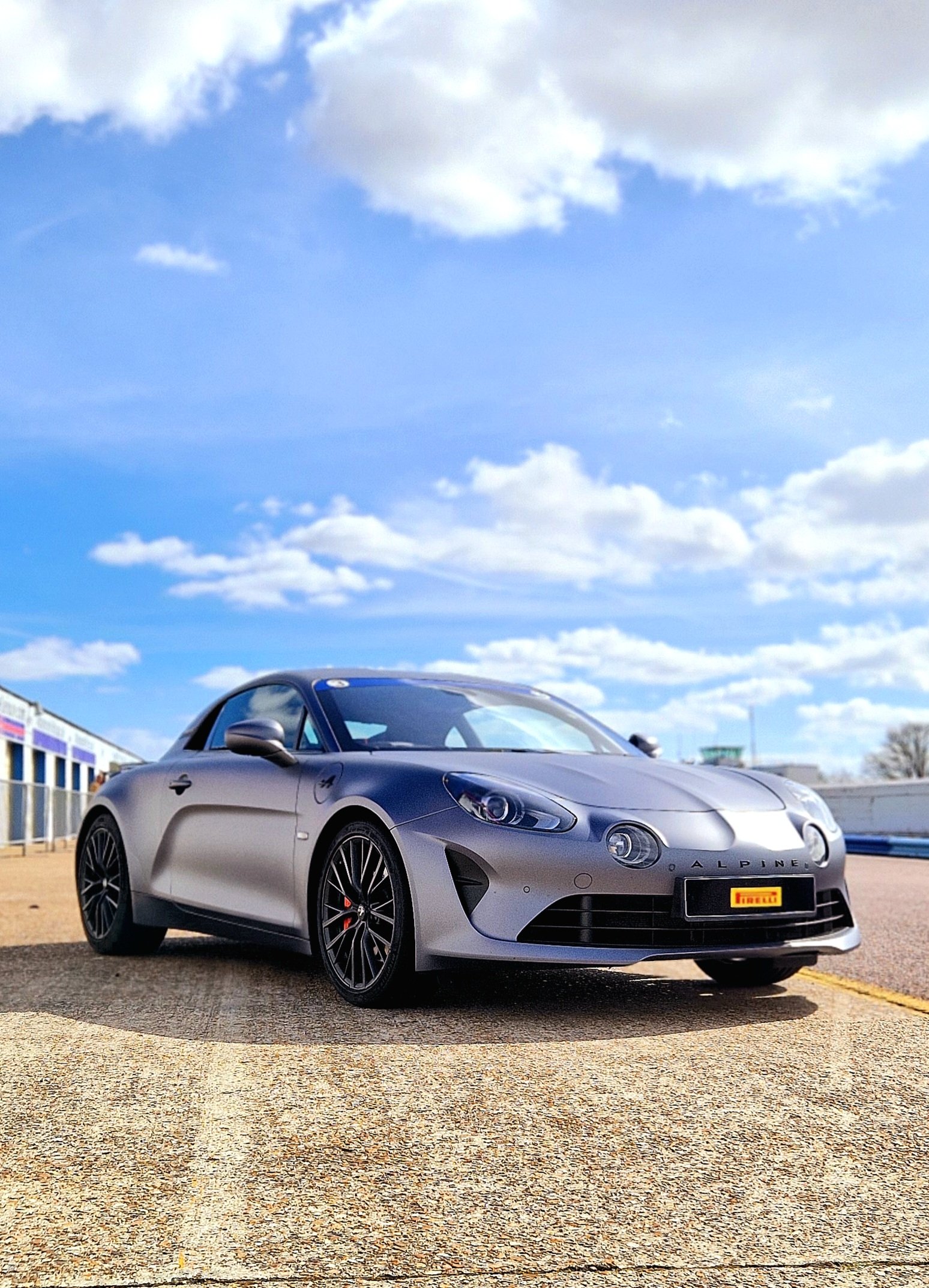 A silver sports car with a sleek design parked on a tarmac surface under a partly cloudy sky.