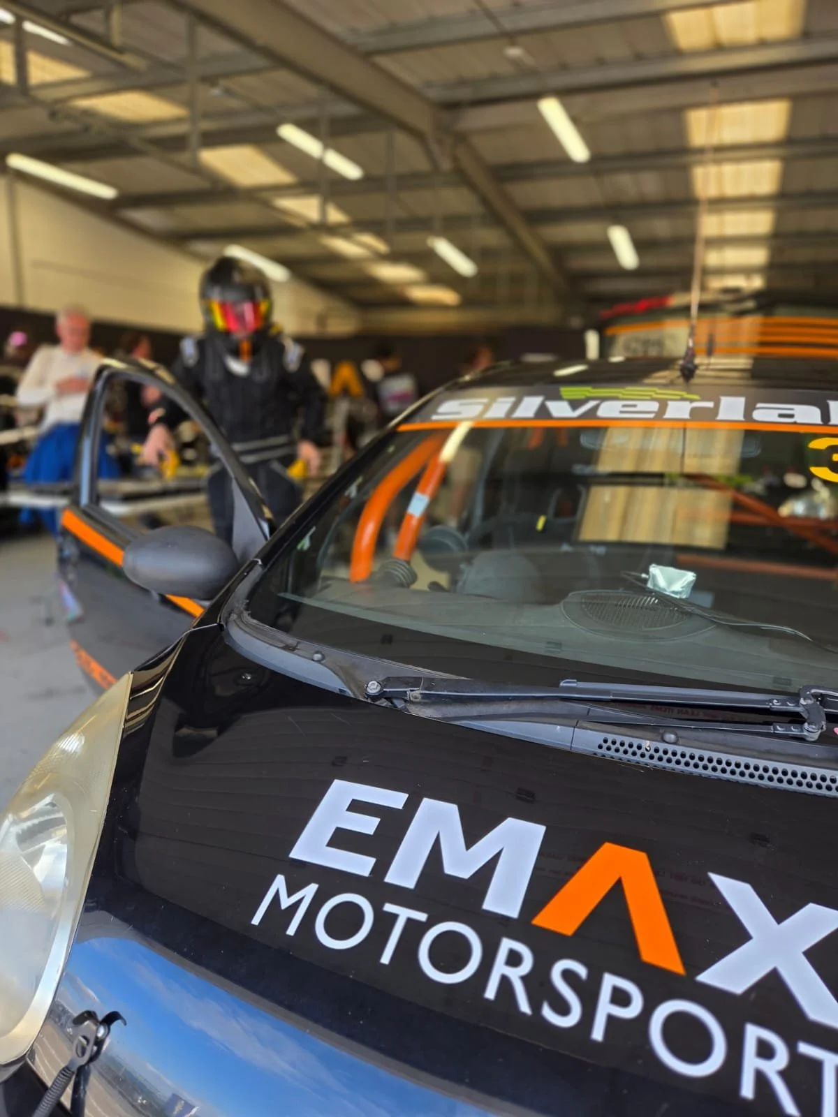 A race car with EMAX Motorsports branding in a garage, a person wearing a racing helmet and suit is in the background.