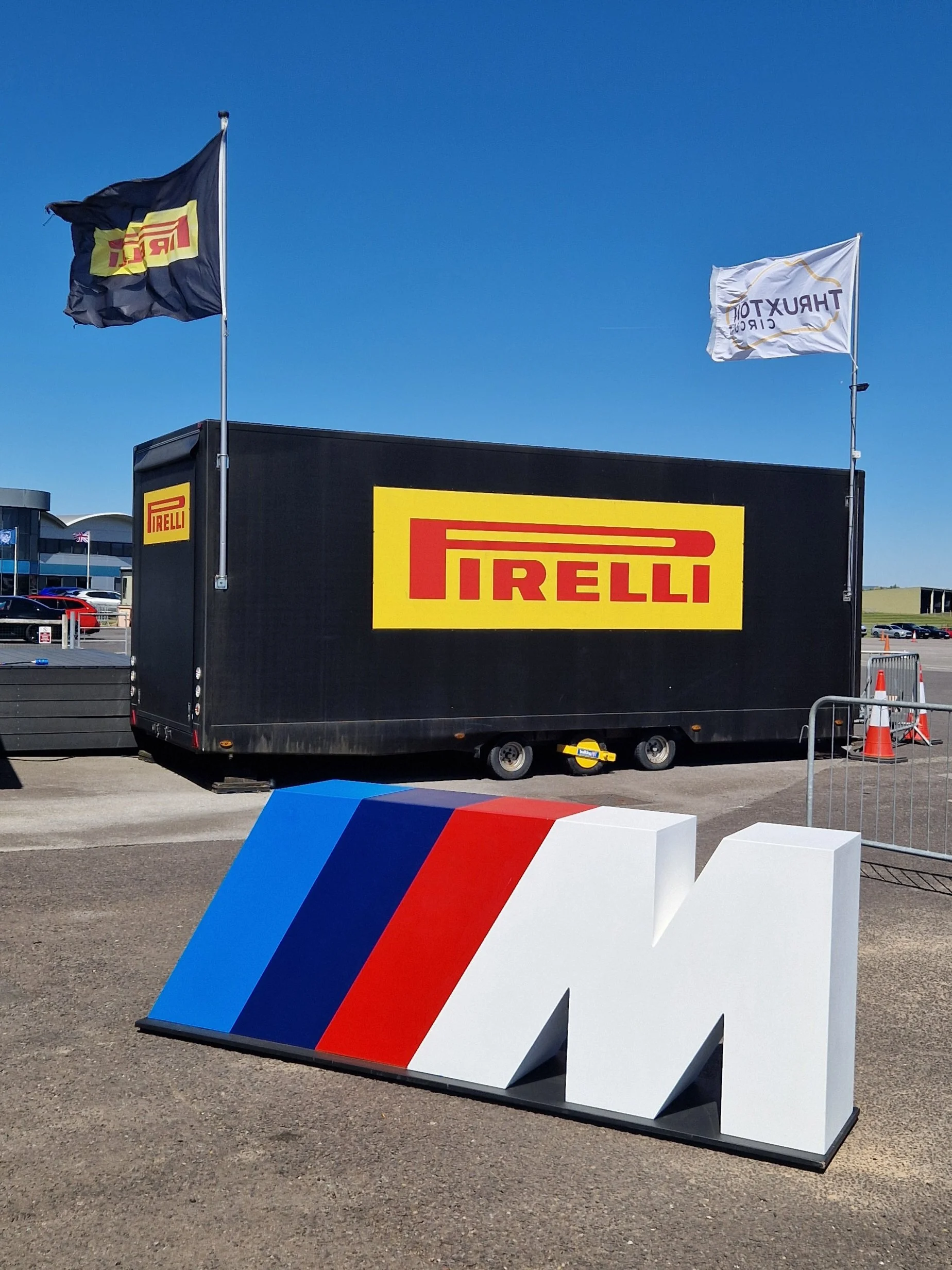 Large black trailer with Pirelli logo, two flags on poles, one with Pirelli logo and the other with Thruxton Circuit logo, in a parking lot with clear blue sky and some buildings in the background.