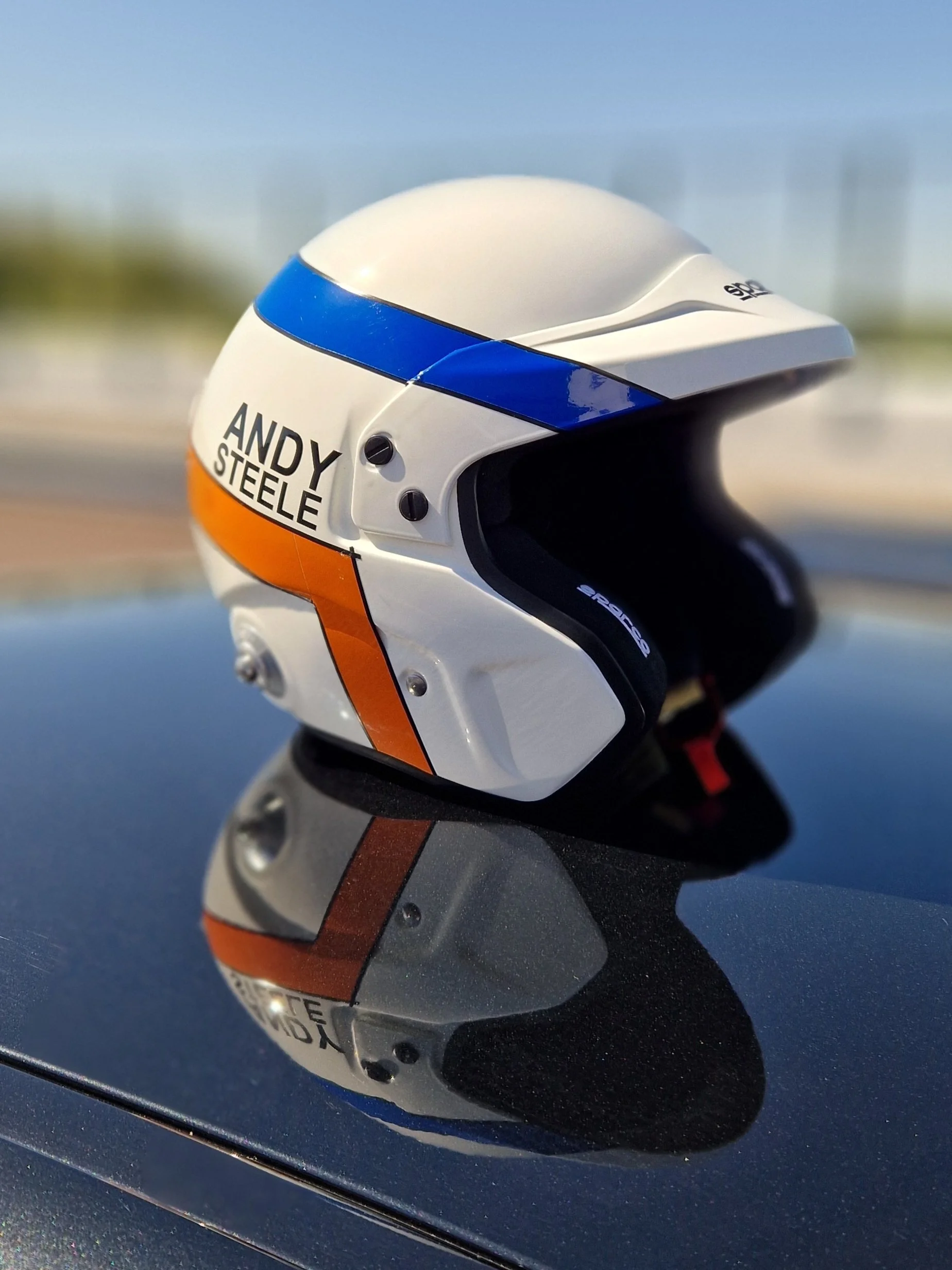 Close-up of a white open face helmet with blue and orange stripes and the name "Andy Steele" printed on the side, reflected on a shiny surface, with a blurred background of a blue sky and buildings.