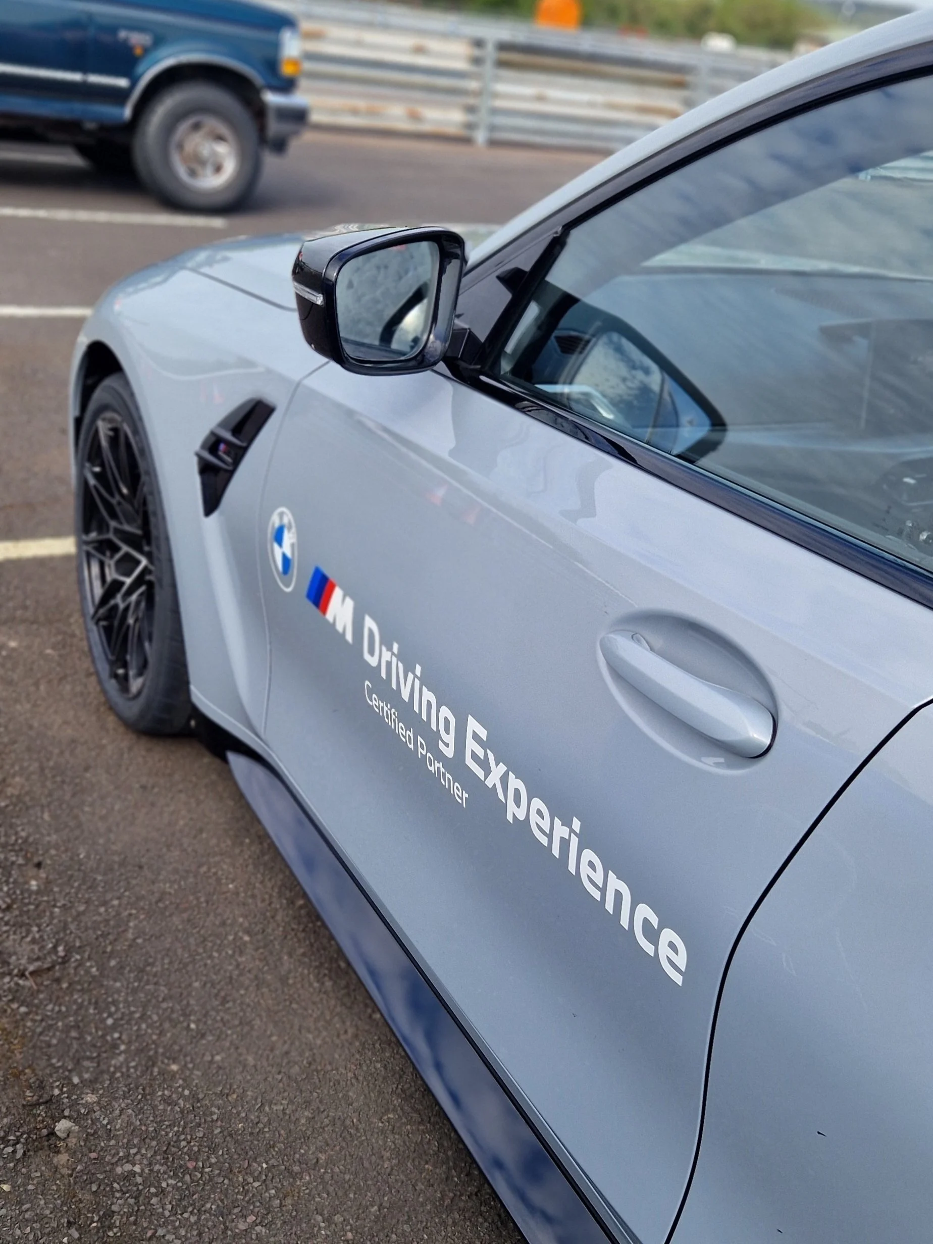 A silver BMW sports car parked on the side of a road with a sign that reads "Driving Excellence, Certified Partner." In the background, there is a blue pickup truck driving on the highway with guardrails and some greenery visible.