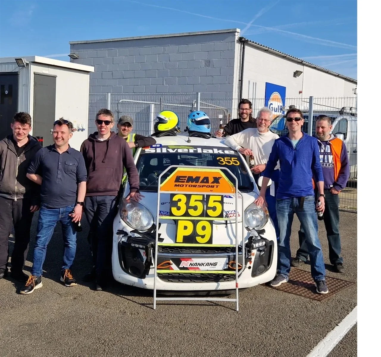Group of people standing around a race car with helmets on top, in an outdoor area with a fence and building in the background, under a clear blue sky.